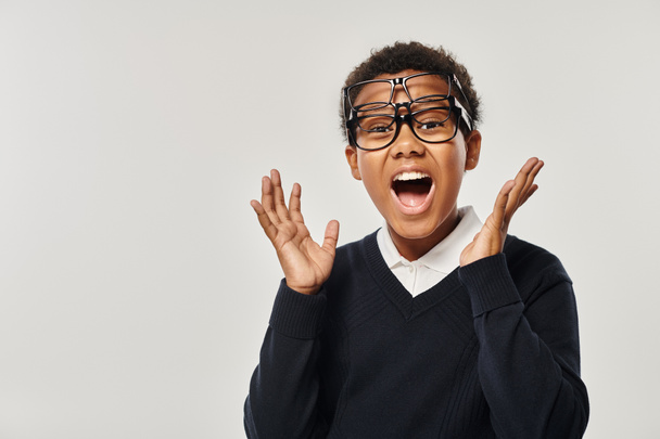 niño afroamericano emocionado en suéter y gafas con gafas y mirando a la cámara en gris - Foto, imagen