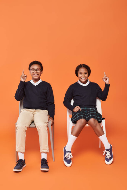 happy african american schoolkids in uniform pointing up and sitting on chairs on orange backdrop - Photo, Image