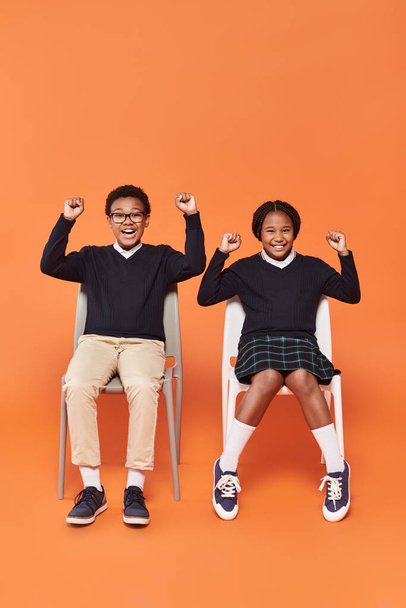 excited african american schoolkids in uniform smiling and sitting on chairs on orange backdrop - Photo, Image