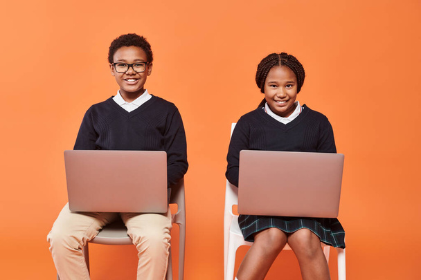 gelukkig Afrikaans amerikaans schoolkinderen in uniform zitten op stoelen en met behulp van laptops op oranje achtergrond - Foto, afbeelding