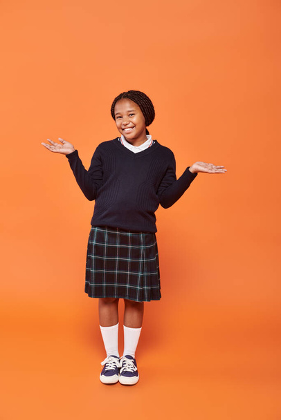 happy african american schoolgirl in uniform gesturing and looking at camera on orange background - Photo, Image