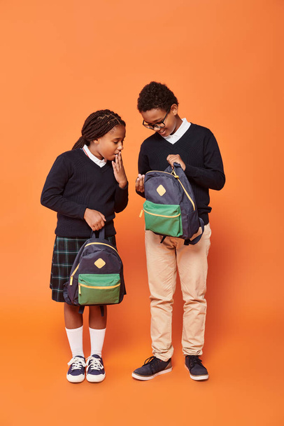 reaction, african american schoolkids in uniform holding backpacks and standing on orange background - Photo, Image