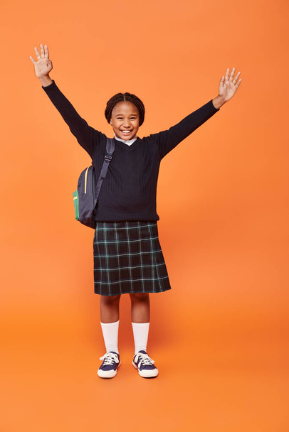 allegra studentessa afroamericana in uniforme sorridente e tenendo lo zaino su sfondo arancione - Foto, immagini