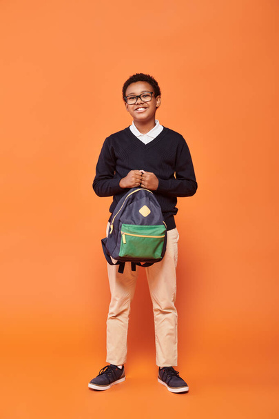 szczęśliwy Afrykański amerykański uczeń w mundurze uśmiecha się i trzyma plecak na pomarańczowym tle - Zdjęcie, obraz