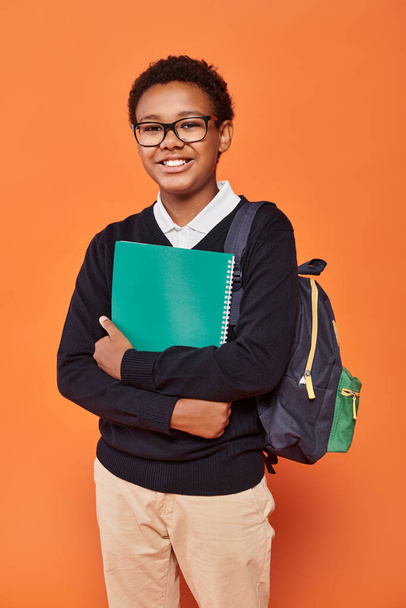 розслаблений афроамериканський школяр в уніформі тримає рюкзак і підручник на помаранчевому тлі - Фото, зображення