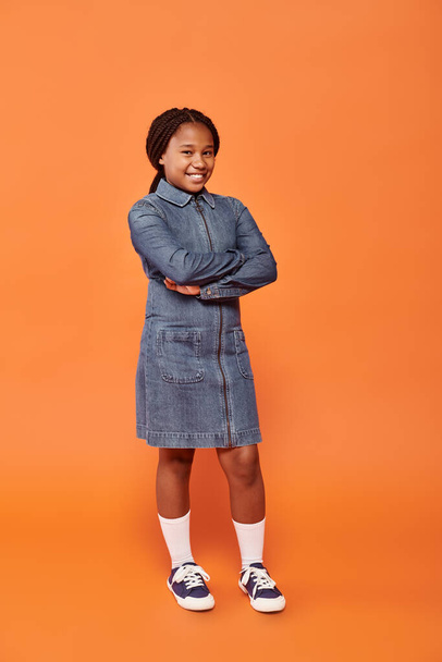 longitud completa de la chica afroamericana feliz en vestido de mezclilla posando con los brazos cruzados sobre fondo naranja - Foto, Imagen