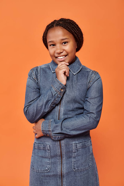 Porträt eines fröhlichen afrikanisch-amerikanischen Mädchens in stylischem Jeanskleid, das auf orangefarbenem Hintergrund lächelt - Foto, Bild