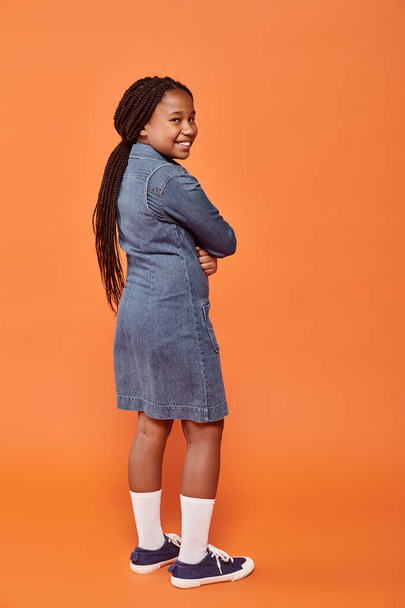 volledige lengte van positief Afrikaans Amerikaans meisje in denim jurk staande op oranje achtergrond - Foto, afbeelding