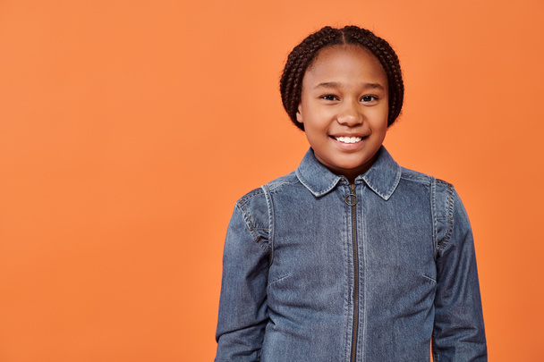 portret van gelukkig Afrikaans Amerikaans meisje met vlechten glimlachen en kijken naar camera op oranje achtergrond - Foto, afbeelding