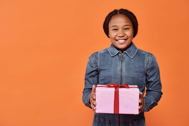 gelukkig afrikaans amerikaans meisje in denim jurk met verpakt cadeau en kijken naar camera op oranje - Foto, afbeelding