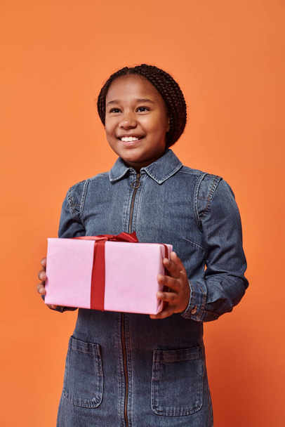 radostná africká americká dívka v džínových šatech drží zabalený dárek a dívá se na kameru na oranžové - Fotografie, Obrázek