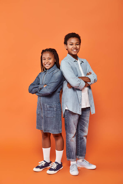 щасливі афроамериканські діти в повсякденному одязі з джинсами, що стоять зі складеними руками на помаранчевому тлі - Фото, зображення