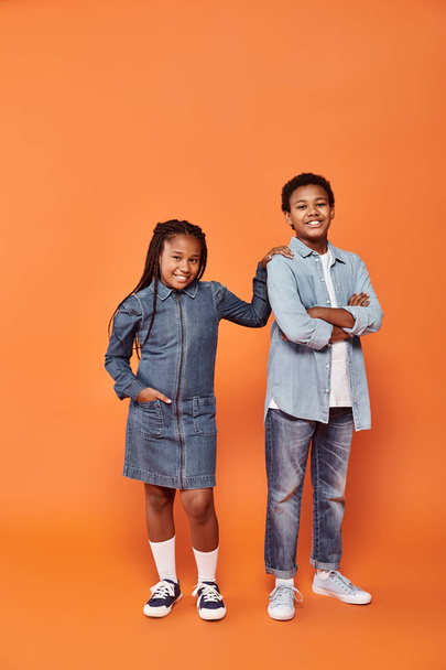 crianças americanas africanas otimistas em traje casual de ganga posando juntas em fundo laranja - Foto, Imagem