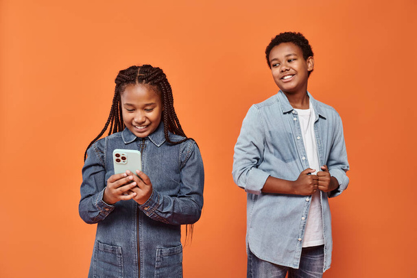 heureuse fille afro-américaine avec des tresses en denim robe en utilisant smartphone près du garçon sur fond orange - Photo, image