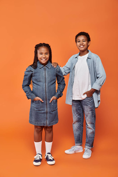 happy african american girl in casual denim attire posing together with boy on orange, full length - Photo, Image