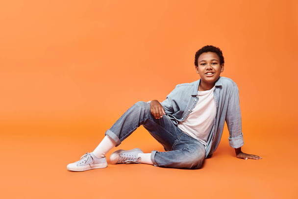 glücklicher afrikanischer amerikanischer Junge in Alltagskleidung posiert auf dem Boden vor orangefarbenem Hintergrund und lächelt in die Kamera - Foto, Bild