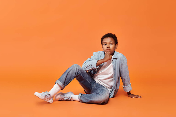pensive preteen african american boy in casual attire sitting on floor and looking at camera - Photo, Image