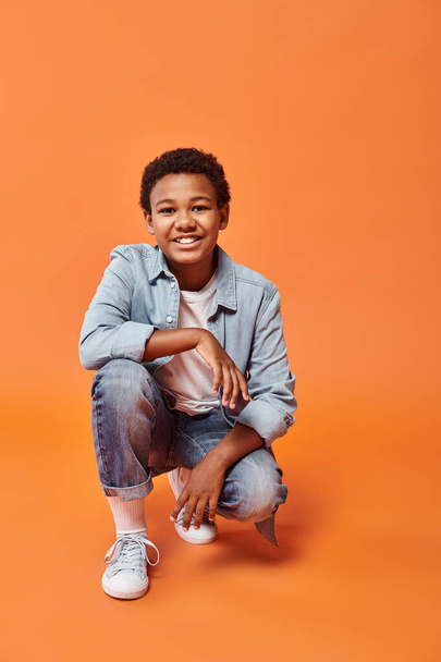 fröhlicher vorpubertärer afrikanisch-amerikanischer Junge in Alltagskleidung, der auf einem Knie vor orangefarbenem Hintergrund steht - Foto, Bild
