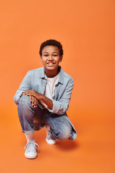 cheerful preteen african american boy in casual outfit standing on one jnee and smiling at camera - Photo, Image
