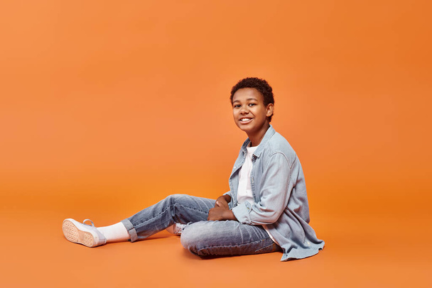 alegre menino afro-americano em traje casual sentado no chão e sorrindo alegremente para a câmera - Foto, Imagem