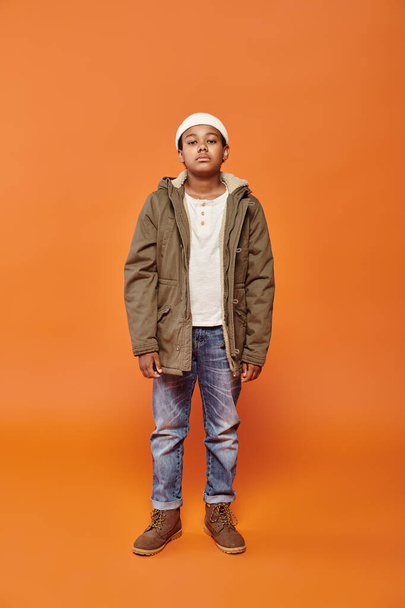 preadolescent african american boy in winter attire with beanie hat posing on orange backdrop - Photo, Image
