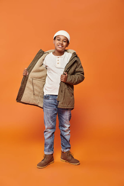 счастливый африканский американский мальчик в зимней теплой куртке позирует на оранжевом фоне и улыбается в камеру - Фото, изображение