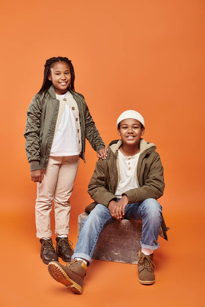 alegre pré-adolescente afro-americano menino e menina em trajes de inverno posando em pano de fundo laranja - Foto, Imagem