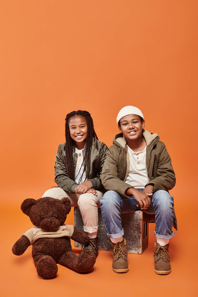 オレンジ色の背景にテディベアを着た暖かい衣装で陽気なアフリカ系アメリカ人の子供たち - 写真・画像