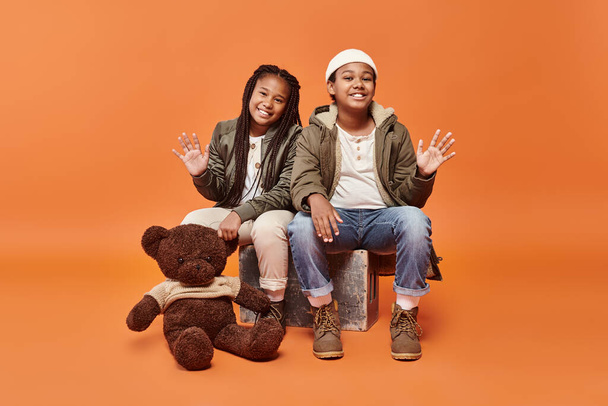 jolly preteen african american children in winter outfit posing with teddy bear and waving at camera - Photo, Image