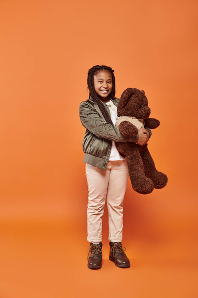 fröhliches afrikanisch-amerikanisches Mädchen mit Zöpfen in Winterkleidung posiert freudig mit ihrem Teddybär - Foto, Bild