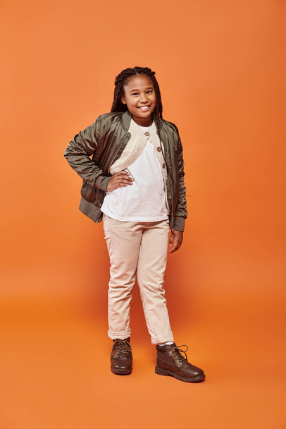 alegre pré-adolescente afro-americana menina com tranças posando com um braço akimbo e sorrindo para a câmera - Foto, Imagem