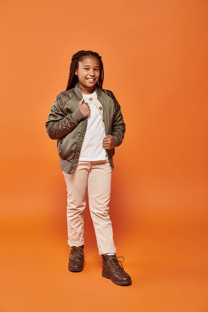 jolly tiener afrikaans amerikaans meisje in warme outfit poseren op oranje achtergrond en glimlachen op camera - Foto, afbeelding