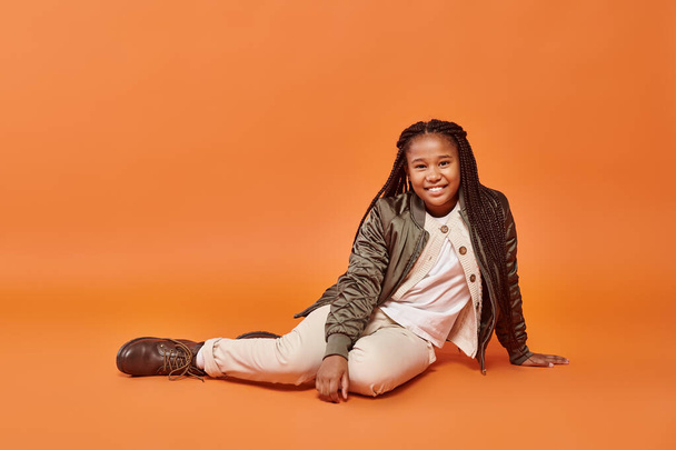cheerful preteen african american girl in warm winter attire sitting on floor and smiling at camera - Photo, Image
