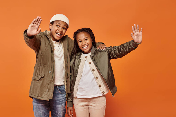 счастливая африканская американская девушка и мальчик в зимних нарядах радостно позируют вместе на оранжевом фоне - Фото, изображение