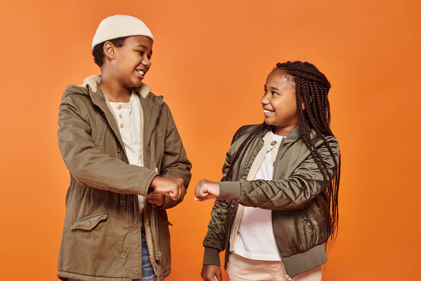 オレンジ色の背景に彼らの拳をぶつける暖かい冬の服装で陽気なアフリカ系アメリカ人の子供 - 写真・画像