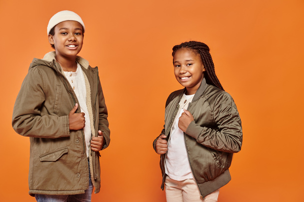 щасливий афроамериканський хлопчик і дівчинка в зимових нарядах посміхаються на камеру на помаранчевому фоні - Фото, зображення
