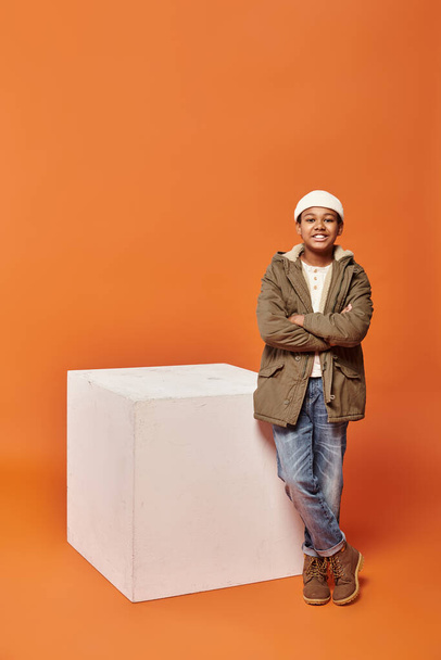 jolly preteen african american boy in warm winter jacket posing near white cube on orange backdrop - Photo, Image
