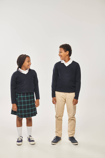 cheerful african american schoolchildren in uniform smiling at each other on gray background - Photo, Image