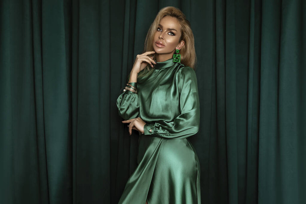 Элегантная молодая женщина в атласно-зеленом платье и украшения на зеленом бархатном фоне в студии. - Фото, изображение