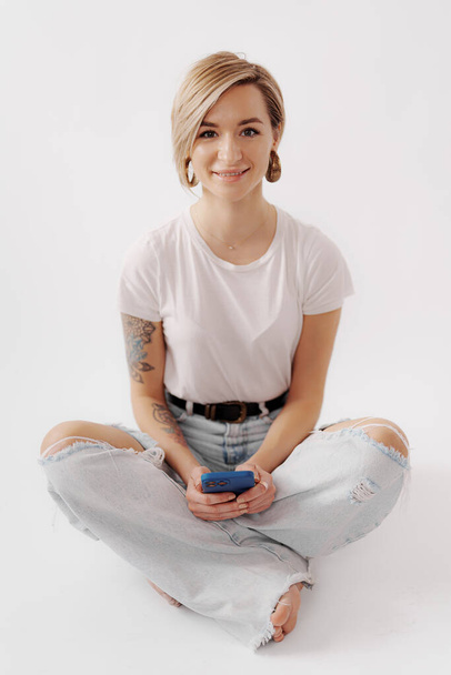 Eine junge Frau mit einem angenehmen Lächeln sitzt im Schneidersitz, während sie ein Telefon in der Hand hält, in einer hellen und luftigen Umgebung - Foto, Bild