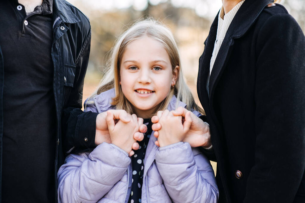 Happy Child Holding Hands with Parents Outdoors. Een lachend jong meisje dat haar ouders handen aan beide kanten vasthoudt, buiten met een gevoel van geborgenheid en liefde. kinderen opgevoed door zowel moeder als vader. - Foto, afbeelding