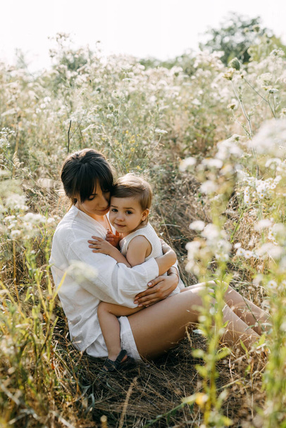 Madre abrazando a la pequeña hija, sonriendo, sentada en un campo con hierba alta. - Foto, Imagen