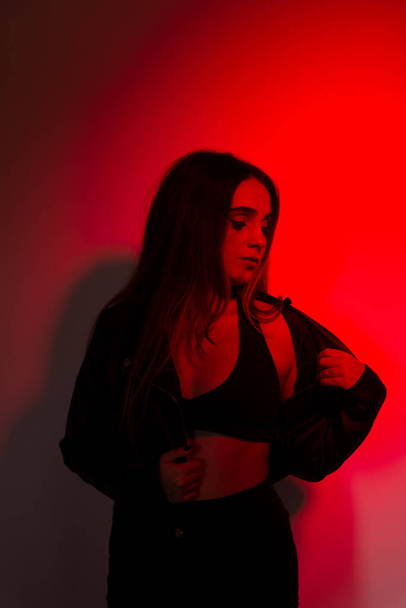 Mooi jong model met zwarte kleding poserend voor foto verlicht in rode kleur. Studio portret - Foto, afbeelding