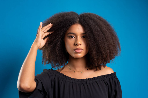Siyah güç saçlarına dokunan güzel bir genç kadının yarı kapalı portresi. Mavi arkaplanda izole. - Fotoğraf, Görsel