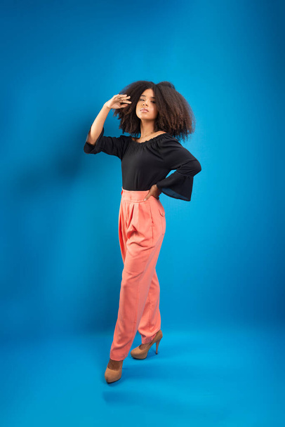 Beautiful young woman, standing, in casual clothes, touching her black power hair. Isolated on blue background. - Photo, Image
