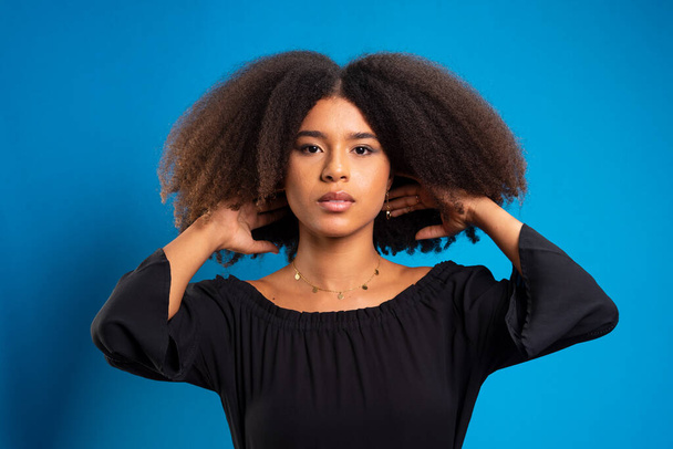 Half-closed portrait of beautiful young woman touching black power hair. Isolated on blue background. - Zdjęcie, obraz