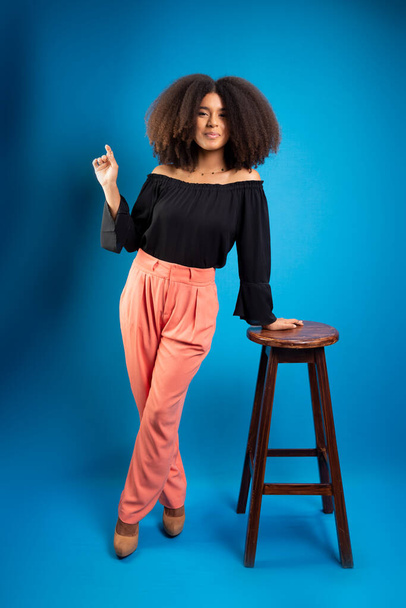 Mladá žena s černými silnými vlasy, opírající se o dřevěnou stoličku ukazující vzhůru. Izolováno na modrém pozadí. - Fotografie, Obrázek