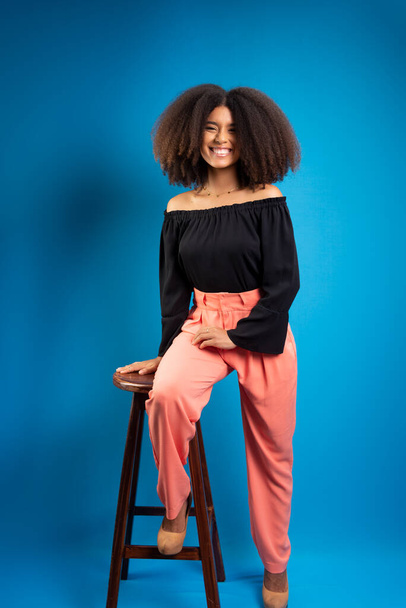 Młoda kobieta z czarnymi włosami, opierająca się na drewnianym stołku, pozująca do zdjęcia. Izolacja na niebieskim tle. - Zdjęcie, obraz