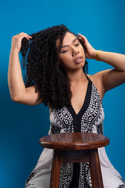 Schöne junge Frau mit lockigem Haar lehnt auf einem Holzhocker und posiert für ein Foto. Vereinzelt auf blauem Hintergrund. - Foto, Bild