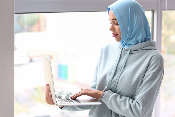 Κομψό μουσουλμάνα γυναίκα χρησιμοποιώντας φορητό υπολογιστή κοντά στο παράθυρο στο σπίτι - Φωτογραφία, εικόνα